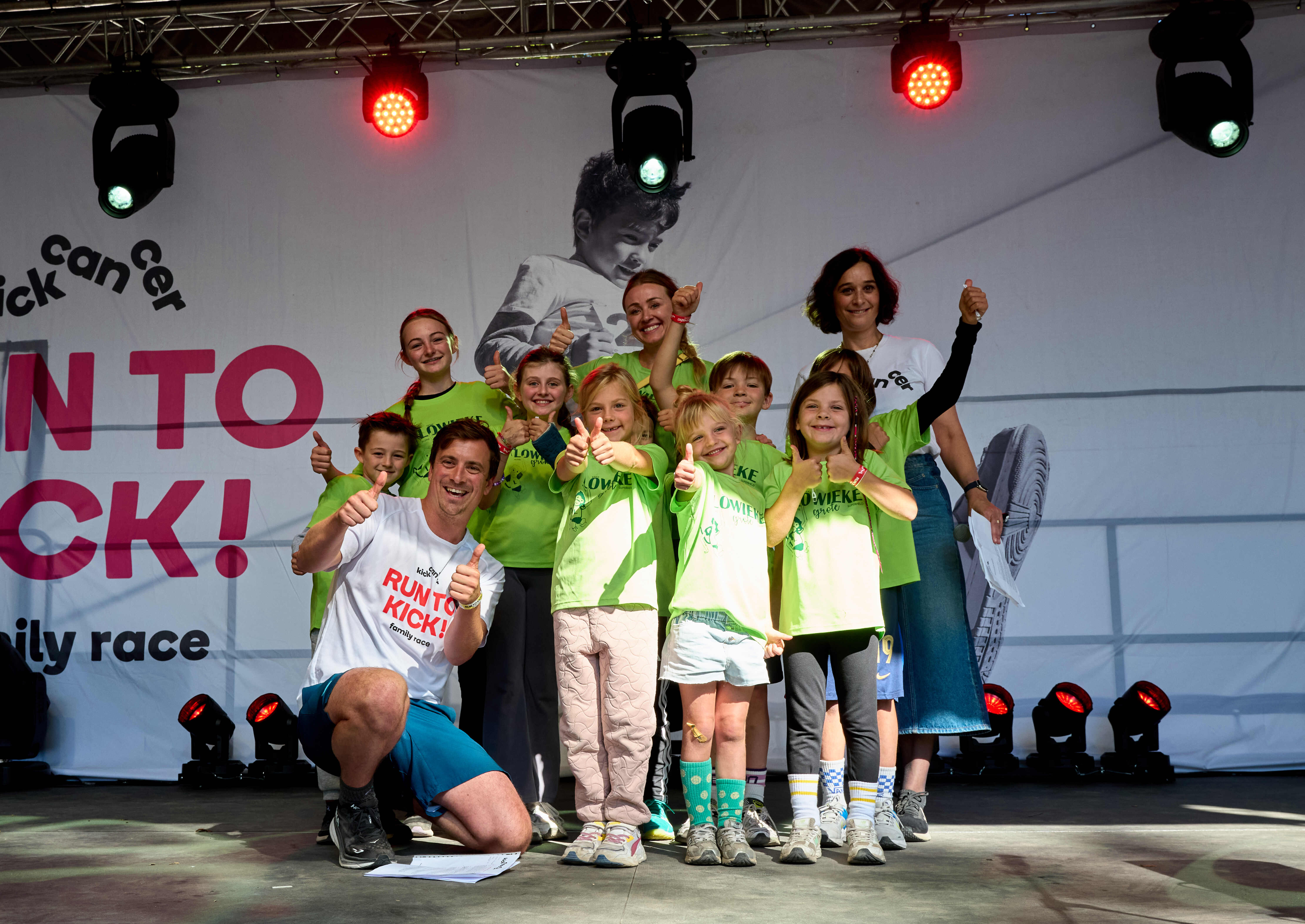 Des enfants et des bénévoles posent sur scène avec l’ambassadeur Niels Destadsbader lors de RUN TO KICK, souriants et levant le pouce pendant l’événement solidaire de KickCancer.