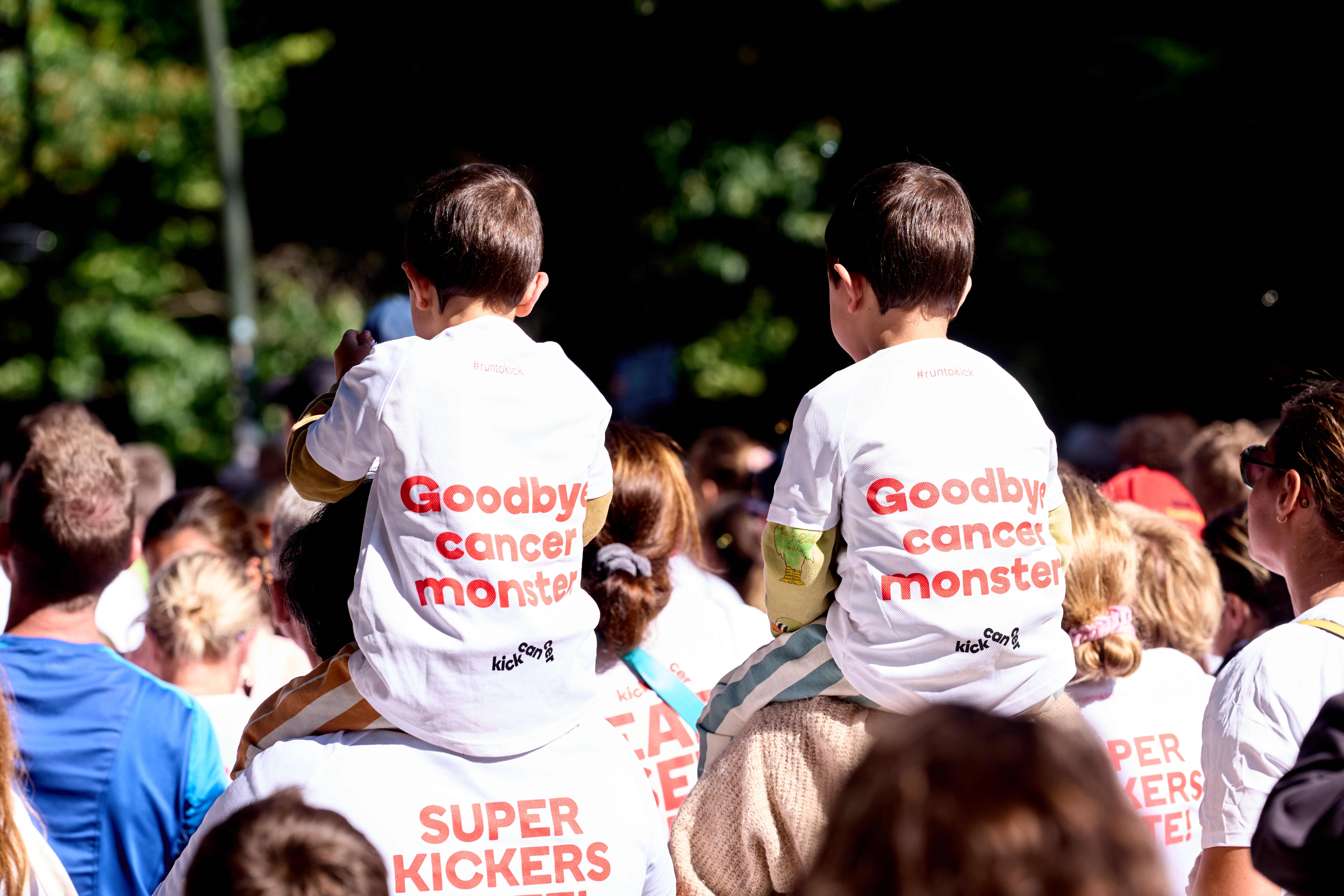 Deux enfants assis sur les épaules d’adultes portent des T-shirts RUN TO KICK avec le message « Goodbye cancer monster » lors de l’événement solidaire de KickCancer.