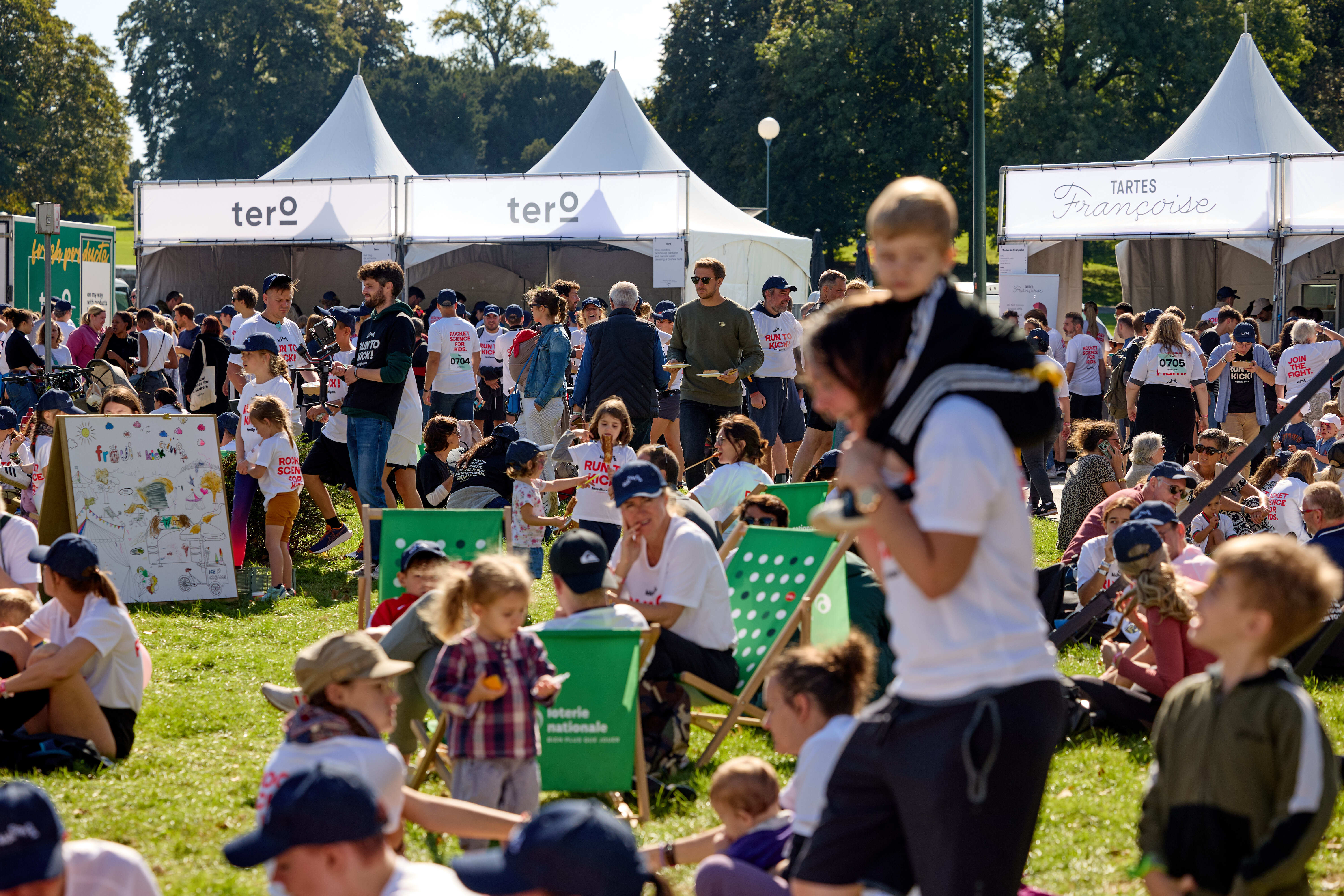 Des familles et des participants se détendent sur l’herbe lors de RUN TO KICK, avec des stands de nourriture et des activités en arrière-plan pendant l’événement solidaire de KickCancer.