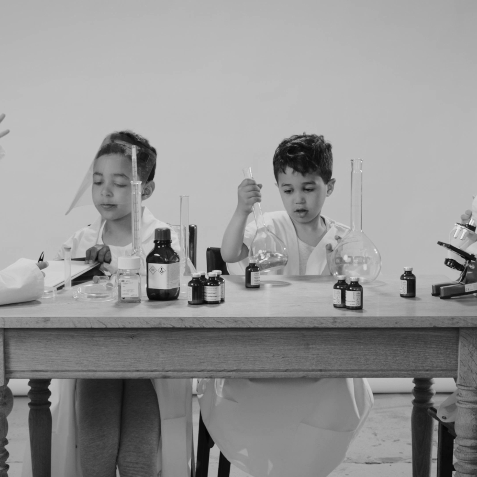 Four young children dressed as scientists sit at a lab table, experimenting with beakers, liquids, and a microscope.
