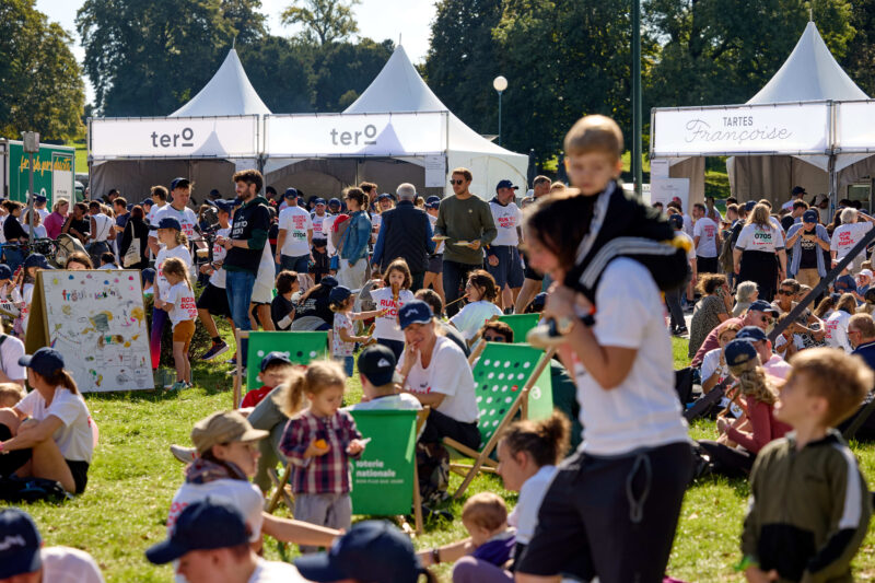 Des familles et des participants se détendent sur l’herbe lors de RUN TO KICK, avec des stands de nourriture et des activités en arrière-plan pendant l’événement solidaire de KickCancer.
