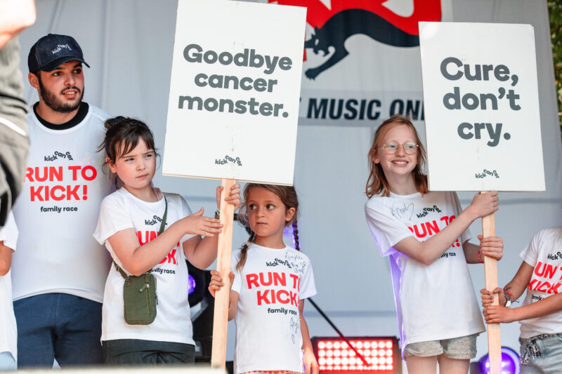 Des enfants portant des T-shirts RUN TO KICK tiennent des pancartes avec les messages « Goodbye cancer monster » et « Cure, don’t cry » sur scène lors de l’événement solidaire de KickCancer.