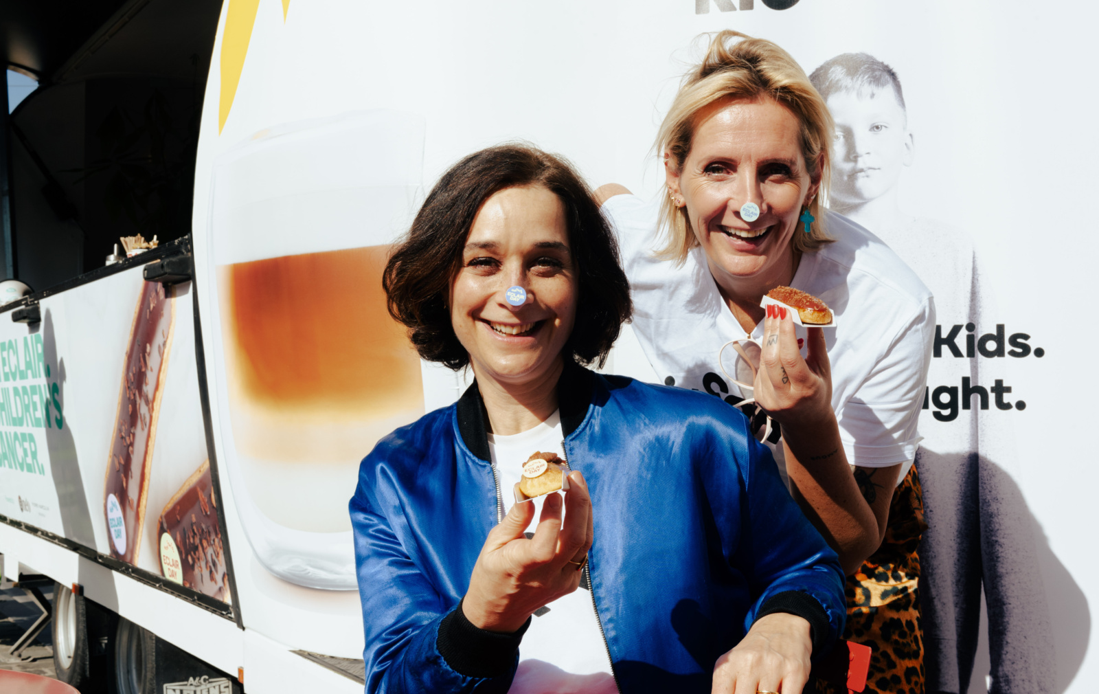 Delphine Heenen et Sofie Dumont, un autocollant Eclair Day sur le nez, brandissent un éclair en riant devant le food truck KickCancer.