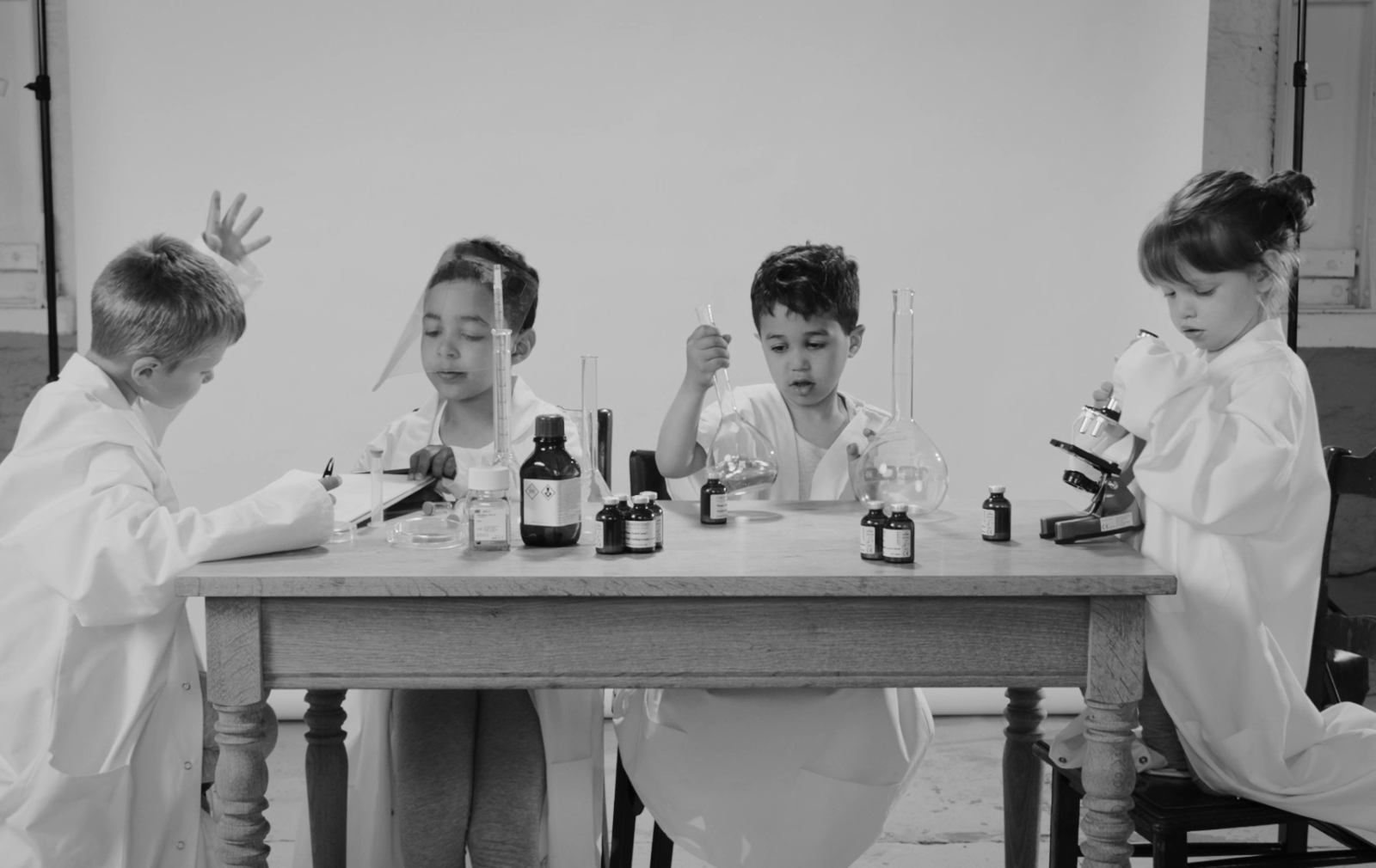 Four young children dressed as scientists sit at a lab table, experimenting with beakers, liquids, and a microscope.